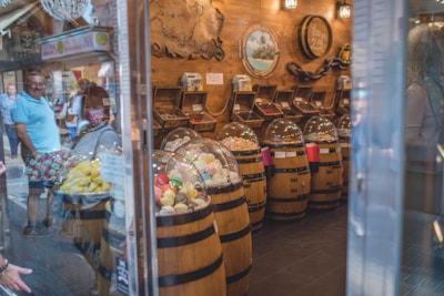 A quaint confectionery store features wooden barrels filled with brightly colored candies, covered with clear dome lids. The barrels are neatly arranged along the store with shelves in the background holding assorted sweets. The warm, wooden interior is adorned with decorative pieces like maps and paintings. Reflections of people observing the display can be seen through the glass exterior.