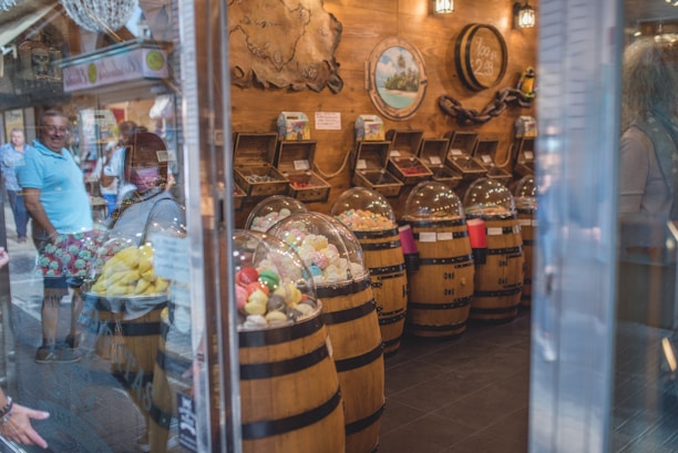 A quaint confectionery store features wooden barrels filled with brightly colored candies, covered with clear dome lids. The barrels are neatly arranged along the store with shelves in the background holding assorted sweets. The warm, wooden interior is adorned with decorative pieces like maps and paintings. Reflections of people observing the display can be seen through the glass exterior.