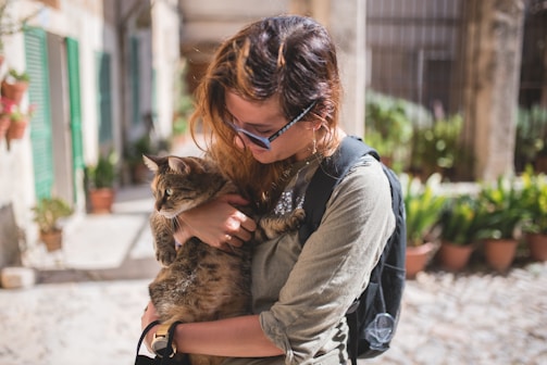 A person wearing sunglasses holds a cat close while standing in an outdoor setting that includes potted plants, stone pathways, and green window shutters. The person has a backpack and seems to be gently cuddling the cat.