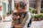 A person wearing sunglasses holds a cat close while standing in an outdoor setting that includes potted plants, stone pathways, and green window shutters. The person has a backpack and seems to be gently cuddling the cat.