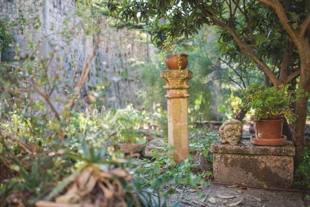 A lush garden scene with a stone pedestal in the center holding a terra-cotta pot with a plant. Surrounding this are various vibrant green plants and trees, creating a serene and natural atmosphere. A carved stone sculpture resembling a face rests on a block next to another pot of foliage.