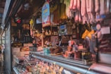 A bustling deli or market stall with an array of cured meats hanging overhead. The counter is filled with a variety of cheeses and meats, while shelves in the background store jars and condiments. The space is dimly lit, creating a cozy and inviting atmosphere.