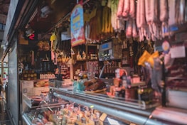 A bustling deli or market stall with an array of cured meats hanging overhead. The counter is filled with a variety of cheeses and meats, while shelves in the background store jars and condiments. The space is dimly lit, creating a cozy and inviting atmosphere.