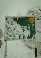 A snow-covered sign stands by the roadside. The sign features a partially visible symbol and some text in Hindi, obscured by thick layers of snow. Ice crystals cling to the edges of the sign, with more snow covering the ground and surrounding fence. Leafless branches and barbed wire fence show signs of accumulation from the heavy snowfall.