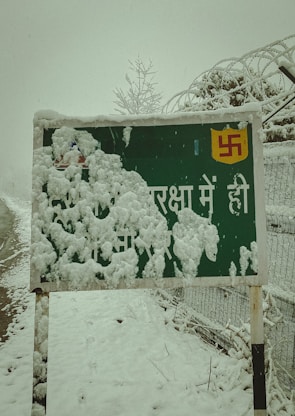 A snow-covered sign stands by the roadside. The sign features a partially visible symbol and some text in Hindi, obscured by thick layers of snow. Ice crystals cling to the edges of the sign, with more snow covering the ground and surrounding fence. Leafless branches and barbed wire fence show signs of accumulation from the heavy snowfall.