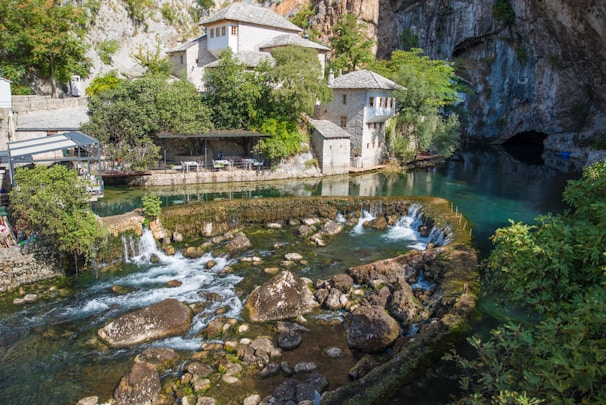 A serene riverside scene featuring historic stone buildings surrounded by lush vegetation. The river flows gently over rocks, creating small waterfalls and leading to a tranquil pool. The architecture blends harmoniously with the natural landscape, featuring traditional stone design and wooden elements, while a cave entrance is visible in the background.