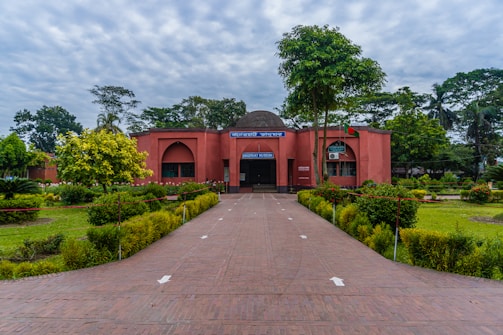 A vibrant pathway leads to a red brick building surrounded by well-maintained gardens with lush green plants and trees. The sky is overcast with clouds creating a serene atmosphere. Flags are visible near the entrance of the building.