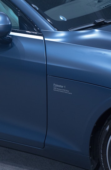 A close-up view of the side of a sleek, blue car displaying the Polestar 1 logo and marking. The vehicle features a high gloss finish, showcasing refined details such as the side mirror and part of the wheel.