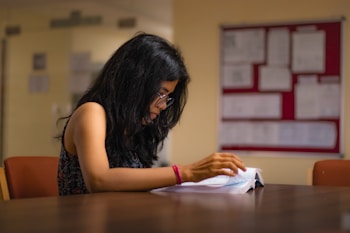 A woman with long black hair and glasses is sitting at a wooden table, deeply engaged in reading a book. The room appears to be a study or library with papers pinned on a board in the background.