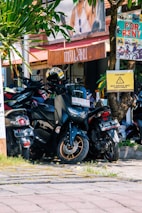 A cluster of motorbikes parked closely together on a street under the shade of a tree. Several signs around indicate the availability of motorbike rentals, with prominent texts like 'For Rent' in bright colors. There is a helmet placed on one of the motorbikes. The setting appears to be in a vibrant area, with greenery and a wooden facade in the background giving a rustic touch.