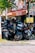 A cluster of motorbikes parked closely together on a street under the shade of a tree. Several signs around indicate the availability of motorbike rentals, with prominent texts like 'For Rent' in bright colors. There is a helmet placed on one of the motorbikes. The setting appears to be in a vibrant area, with greenery and a wooden facade in the background giving a rustic touch.