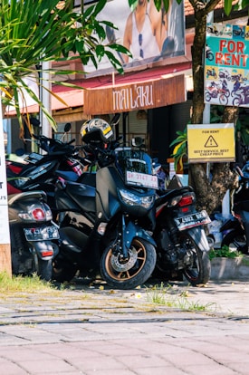 A cluster of motorbikes parked closely together on a street under the shade of a tree. Several signs around indicate the availability of motorbike rentals, with prominent texts like 'For Rent' in bright colors. There is a helmet placed on one of the motorbikes. The setting appears to be in a vibrant area, with greenery and a wooden facade in the background giving a rustic touch.
