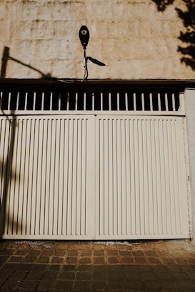 A beige-colored wall with a matching gate featuring vertical slats. Above the gate, there is a mounted black security camera with a visible shadow. The ground is paved with brown tiles, and there are shadows cast by objects not visible in the frame on both the wall and the ground.