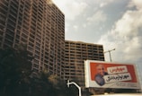 Tall residential buildings with numerous balconies under a partly cloudy sky. In the foreground, a billboard displays a man leaning on an orange background with text in a non-English language.