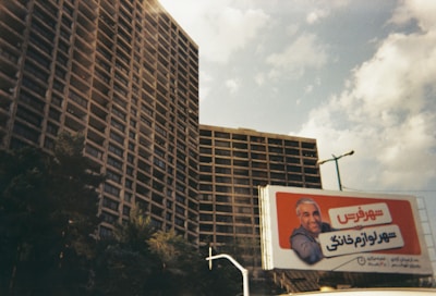 Tall residential buildings with numerous balconies under a partly cloudy sky. In the foreground, a billboard displays a man leaning on an orange background with text in a non-English language.