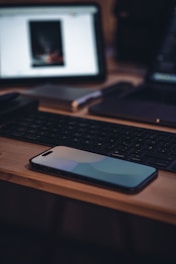 A sleek modern smartphone resting on a wooden desk with a blurred laptop in the background.