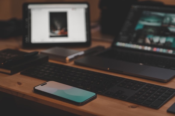 A workspace setup features a smartphone placed on a wooden desk, flanked by a wireless keyboard. In the background, there are two screens, likely a tablet and a laptop, displaying various digital content. A notebook and a pen are partially visible, suggesting a combination of digital and traditional work tools.