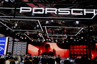 A large crowd gathers at a car exhibition booth prominently featuring the Porsche brand. The space is illuminated with dynamic lighting, including overhead fixtures and vibrant red and black tones. The background features a large screen displaying a silhouette of a person, adding drama to the scene. Reflections and a mix of modern metal textures create an energetic atmosphere.