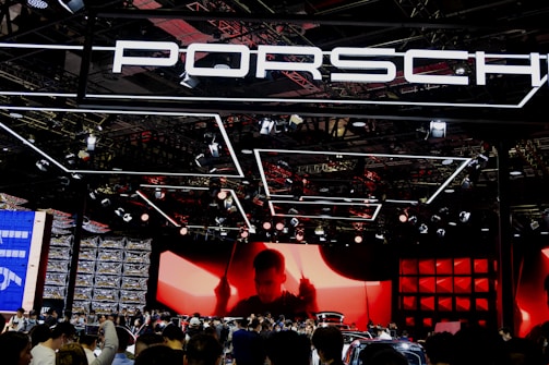 A large crowd gathers at a car exhibition booth prominently featuring the Porsche brand. The space is illuminated with dynamic lighting, including overhead fixtures and vibrant red and black tones. The background features a large screen displaying a silhouette of a person, adding drama to the scene. Reflections and a mix of modern metal textures create an energetic atmosphere.