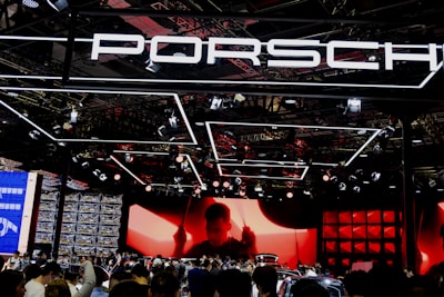 A large crowd gathers at a car exhibition booth prominently featuring the Porsche brand. The space is illuminated with dynamic lighting, including overhead fixtures and vibrant red and black tones. The background features a large screen displaying a silhouette of a person, adding drama to the scene. Reflections and a mix of modern metal textures create an energetic atmosphere.