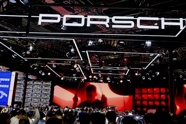 A large crowd gathers at a car exhibition booth prominently featuring the Porsche brand. The space is illuminated with dynamic lighting, including overhead fixtures and vibrant red and black tones. The background features a large screen displaying a silhouette of a person, adding drama to the scene. Reflections and a mix of modern metal textures create an energetic atmosphere.