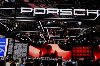A large crowd gathers at a car exhibition booth prominently featuring the Porsche brand. The space is illuminated with dynamic lighting, including overhead fixtures and vibrant red and black tones. The background features a large screen displaying a silhouette of a person, adding drama to the scene. Reflections and a mix of modern metal textures create an energetic atmosphere.