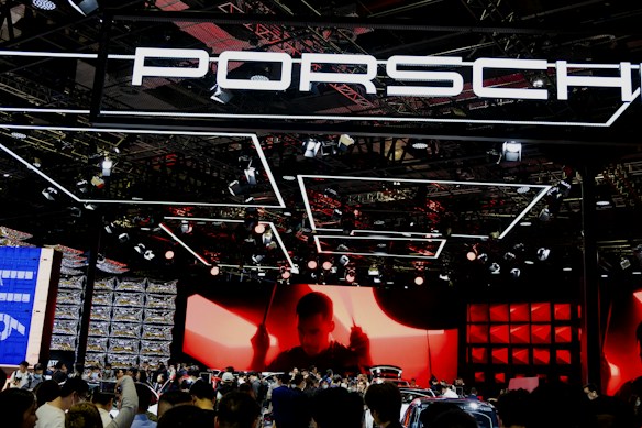 A large crowd gathers at a car exhibition booth prominently featuring the Porsche brand. The space is illuminated with dynamic lighting, including overhead fixtures and vibrant red and black tones. The background features a large screen displaying a silhouette of a person, adding drama to the scene. Reflections and a mix of modern metal textures create an energetic atmosphere.