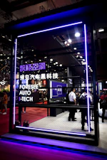 A futuristic exhibit space is enclosed by a mesh fence, highlighted with purple neon lighting. The setting includes a display of technological advancements with several attendees observing the inside. Signage in both English and Chinese indicates a focus on automotive technology.