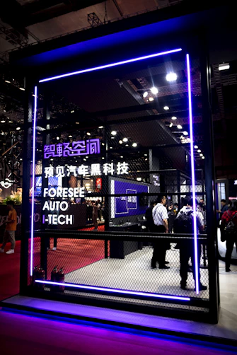 A futuristic exhibit space is enclosed by a mesh fence, highlighted with purple neon lighting. The setting includes a display of technological advancements with several attendees observing the inside. Signage in both English and Chinese indicates a focus on automotive technology.