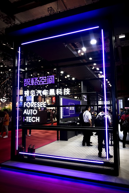 A futuristic exhibit space is enclosed by a mesh fence, highlighted with purple neon lighting. The setting includes a display of technological advancements with several attendees observing the inside. Signage in both English and Chinese indicates a focus on automotive technology.
