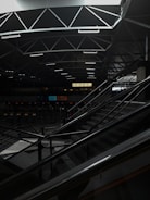 A dimly lit indoor space with modern architectural elements, featuring a geometric ceiling with intersecting lines. An escalator occupies part of the scene, leading up to a higher level. The area is relatively dark, with some areas illuminated by artificial lighting. The sign reading 'EXECUTIVE' is visible in the background.