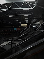 A dimly lit indoor space with modern architectural elements, featuring a geometric ceiling with intersecting lines. An escalator occupies part of the scene, leading up to a higher level. The area is relatively dark, with some areas illuminated by artificial lighting. The sign reading 'EXECUTIVE' is visible in the background.