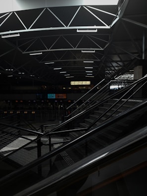 A dimly lit indoor space with modern architectural elements, featuring a geometric ceiling with intersecting lines. An escalator occupies part of the scene, leading up to a higher level. The area is relatively dark, with some areas illuminated by artificial lighting. The sign reading 'EXECUTIVE' is visible in the background.