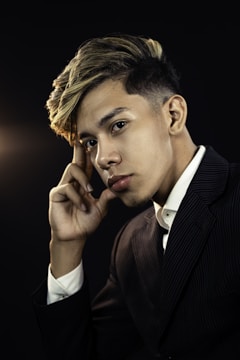 A young person with styled hair and wearing a dark pinstripe suit poses thoughtfully against a dark background. Their expression is contemplative, with one hand resting near their face.