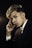 A young person with styled hair and wearing a dark pinstripe suit poses thoughtfully against a dark background. Their expression is contemplative, with one hand resting near their face.