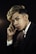 A young person with styled hair and wearing a dark pinstripe suit poses thoughtfully against a dark background. Their expression is contemplative, with one hand resting near their face.