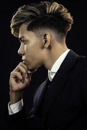 A profile view of a person with styled hair, wearing a dark pinstripe suit and white shirt, resting their chin on their hand in a thoughtful pose against a dark background.