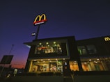 A fast-food restaurant with a prominent red and yellow logo is illuminated during the evening. The building features a glass facade showcasing the interior lighting and activity inside. The sky behind the building is dark with tones of purple and orange indicating dusk.