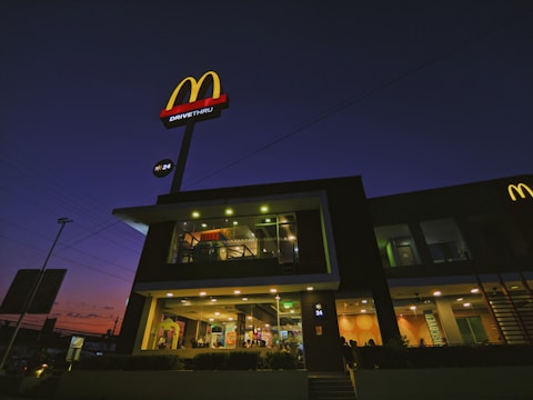 A fast-food restaurant with a prominent red and yellow logo is illuminated during the evening. The building features a glass facade showcasing the interior lighting and activity inside. The sky behind the building is dark with tones of purple and orange indicating dusk.