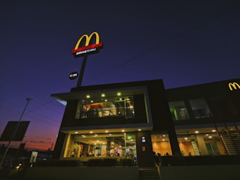A fast-food restaurant with a prominent red and yellow logo is illuminated during the evening. The building features a glass facade showcasing the interior lighting and activity inside. The sky behind the building is dark with tones of purple and orange indicating dusk.