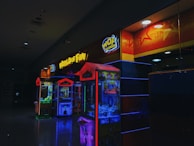A dimly lit arcade with neon lights, featuring claw machines under a sign that says 'World of Fun'. The machines are colorful, displaying bright reds, blues, and yellows.