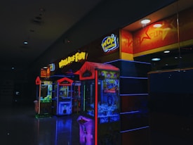 A dimly lit arcade with neon lights, featuring claw machines under a sign that says 'World of Fun'. The machines are colorful, displaying bright reds, blues, and yellows.