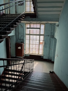 A dimly lit stairwell with rusty metal railings and worn-out steps. The walls are painted in a faded light blue, displaying signs of age and neglect. A large window on the landing allows some natural light to filter in, partially illuminating the area. Electrical wiring is visible along the walls, and a fluorescent light fixture hangs from the ceiling.