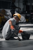 A construction worker wearing a yellow hard hat and orange gloves is squatting on the ground, smoothing concrete with a brush near a black bucket. The worker is dressed in a checkered shirt and has a mask hanging below his chin. Another worker in a straw hat is visible in the background.