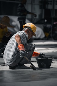 A construction worker wearing a yellow hard hat and orange gloves is squatting on the ground, smoothing concrete with a brush near a black bucket. The worker is dressed in a checkered shirt and has a mask hanging below his chin. Another worker in a straw hat is visible in the background.