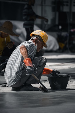 A construction worker wearing a yellow hard hat and orange gloves is squatting on the ground, smoothing concrete with a brush near a black bucket. The worker is dressed in a checkered shirt and has a mask hanging below his chin. Another worker in a straw hat is visible in the background.