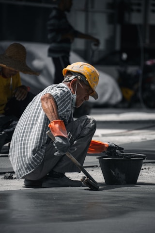 A construction worker wearing a yellow hard hat and orange gloves is squatting on the ground, smoothing concrete with a brush near a black bucket. The worker is dressed in a checkered shirt and has a mask hanging below his chin. Another worker in a straw hat is visible in the background.