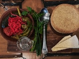 A rustic wooden table hosts a small spread featuring a clay bowl filled with vibrant pickled vegetables, including green peppers, red shredded cabbage, and a whole tomato. Next to the bowl is a spoon and a sprig of fresh parsley. A small glass with clear liquid, likely water or vodka, is situated beside a stack of slices of thin, round flatbread, accompanied by triangular pieces of another type of flatbread.