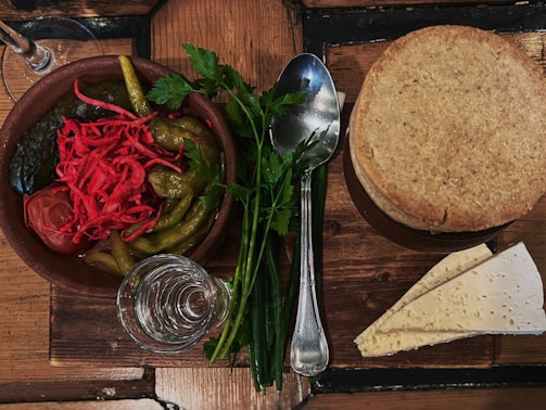 A rustic wooden table hosts a small spread featuring a clay bowl filled with vibrant pickled vegetables, including green peppers, red shredded cabbage, and a whole tomato. Next to the bowl is a spoon and a sprig of fresh parsley. A small glass with clear liquid, likely water or vodka, is situated beside a stack of slices of thin, round flatbread, accompanied by triangular pieces of another type of flatbread.