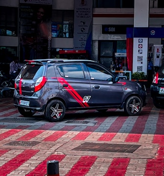 A compact hatchback car with sporty red stripes and the number '07' on its side is parked in a public area with a patterned red and gray tiled floor. The background includes a financial institution, some signage, and partially visible people.
